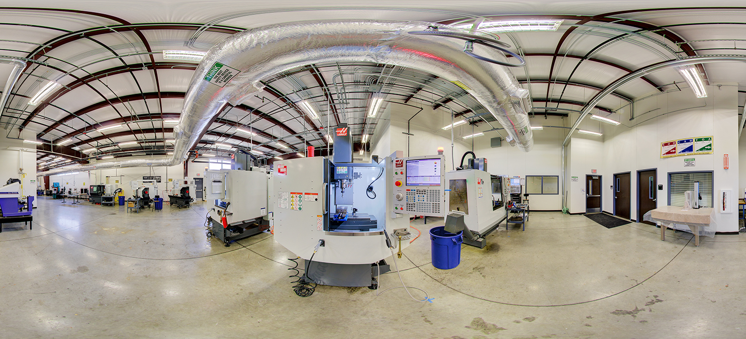 Panoramic view of an industrial machining lab area with multiple CNC machines, control panels, and a large overhead ventilation duct system, surrounded by open floor space and several doors along the walls.