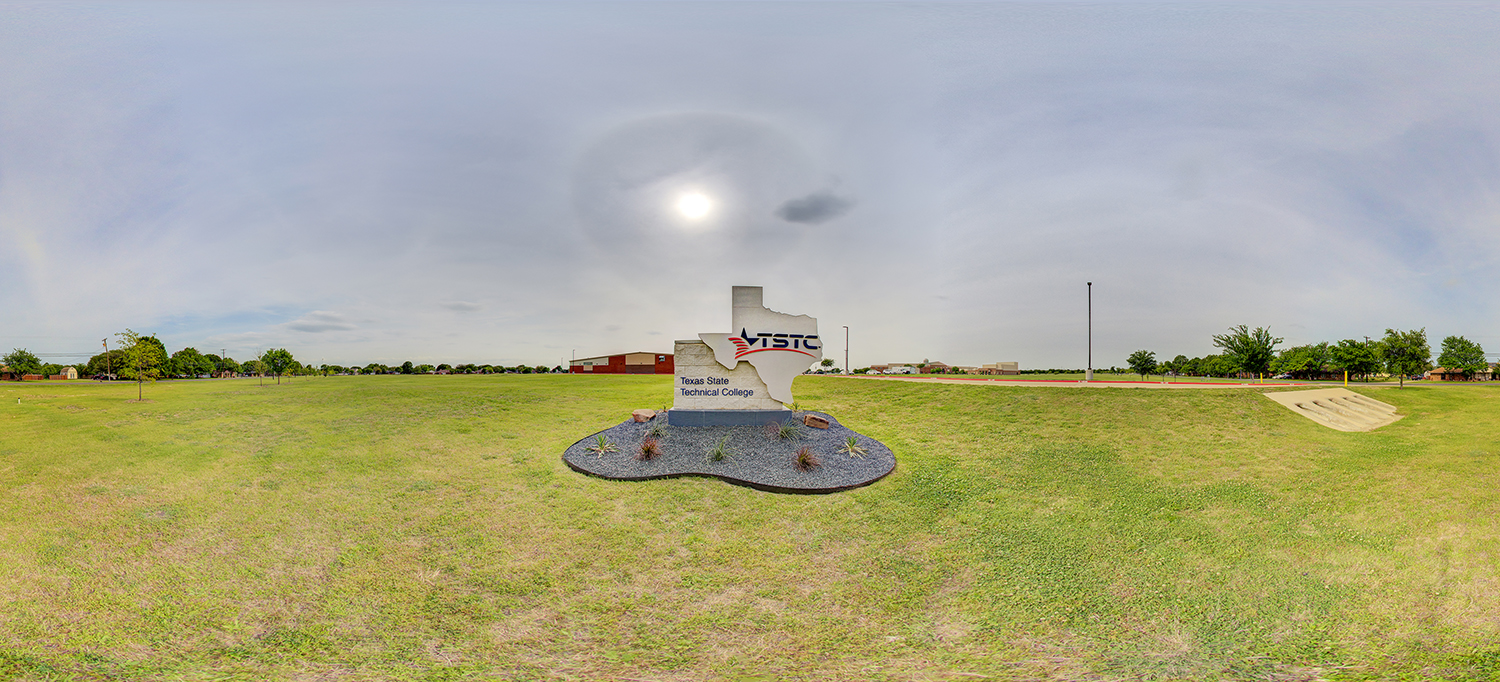 Panoramic view of an outdoor campus area with a Texas-shaped stone sign displaying the TSTC (Texas State Technical College) logo, set in a landscaped patch surrounded by grass, with open fields and buildings in the distance under a bright sky.