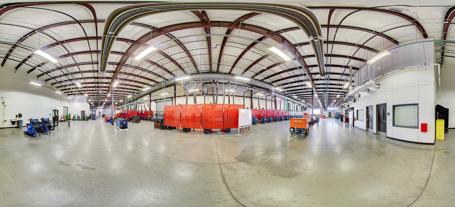 Panoramic view of a large welding lab area with rows of welding stations enclosed by red safety curtains, overhead ventilation ducts, and industrial equipment spread across a wide open concrete floor.