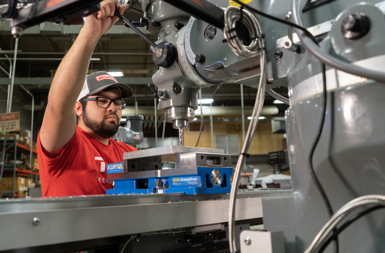 A man in a red shirt and cap operates a milling machine, lowering a drill bit into a metal block secured in a vise inside a workshop.