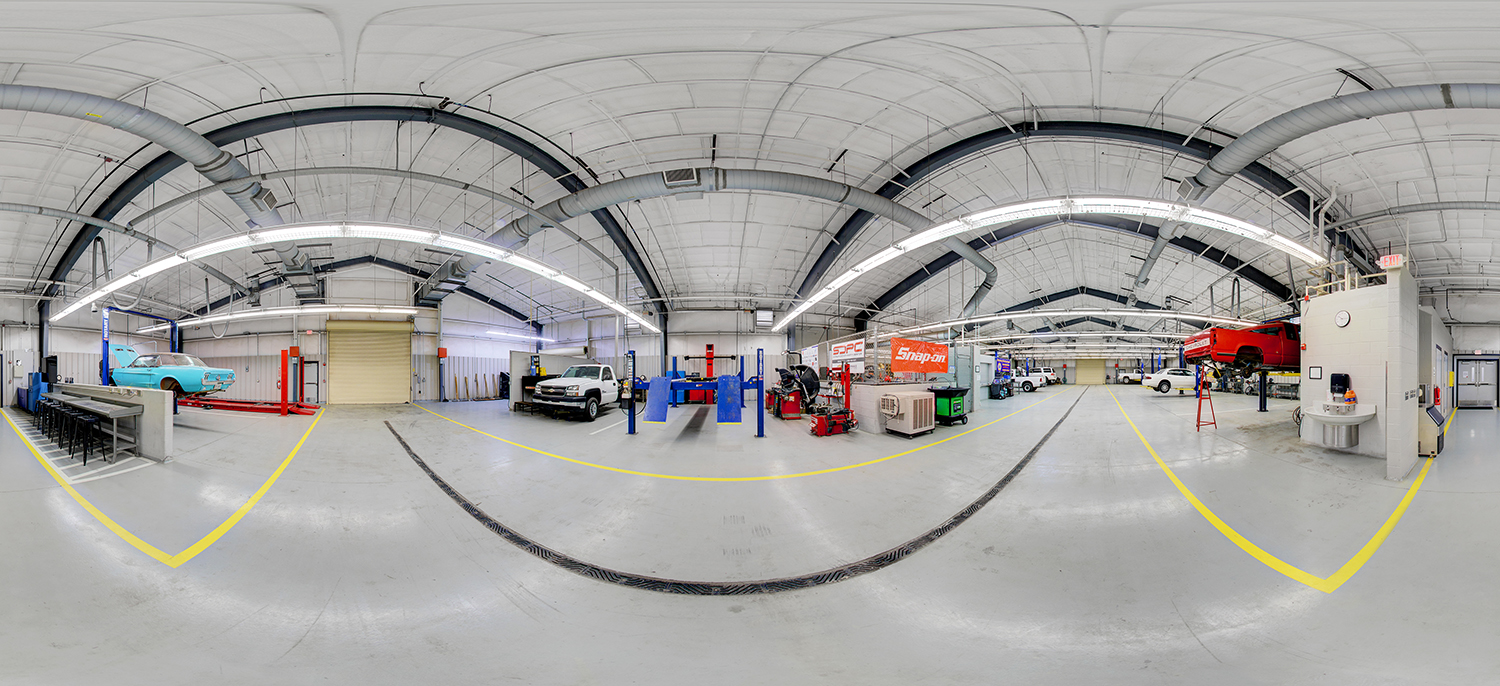 Panoramic view of a large automotive training lab with multiple vehicle lifts, cars and trucks in service bays, tool stations including a Snap-on area, and wide open floor space marked with yellow lines beneath exposed ductwork and bright overhead lighting.