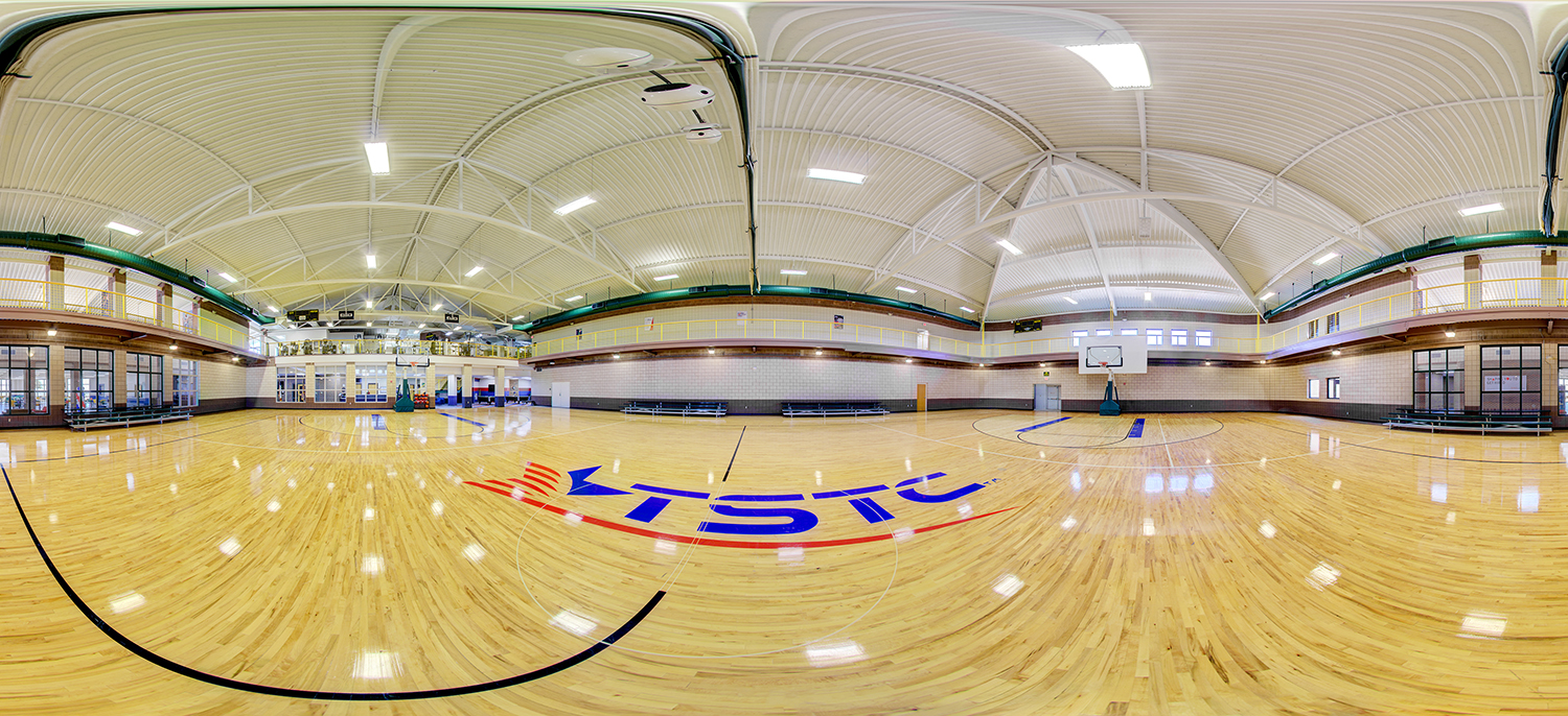 Panoramic view of an indoor gymnasium with a polished wooden basketball court, multiple hoops, bleachers along the walls, and an upper-level track or viewing area beneath a high, arched ceiling with exposed beams.