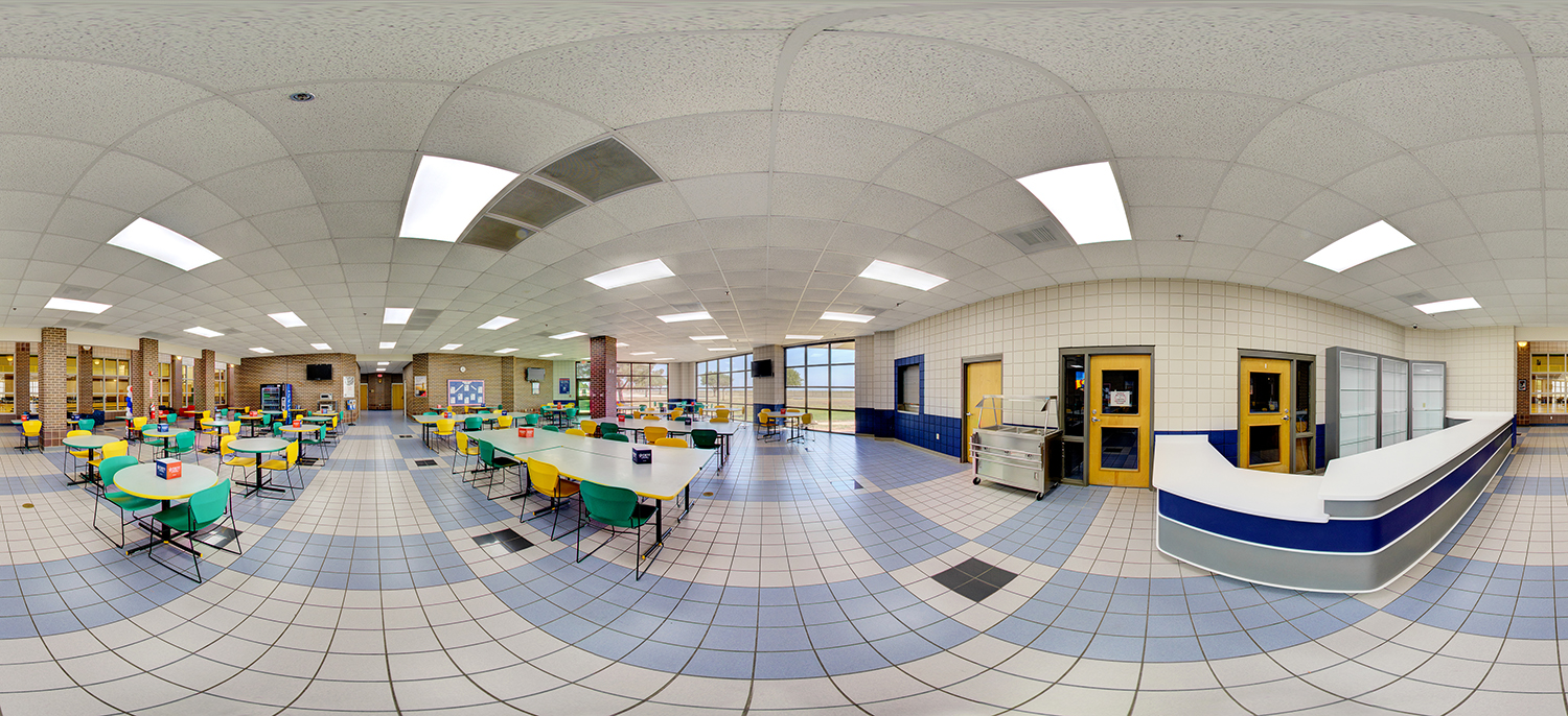 Panoramic view of a bright cafeteria with tiled floors, rows of tables and colorful chairs, large windows letting in natural light, and a serving counter area with doors leading to the kitchen.