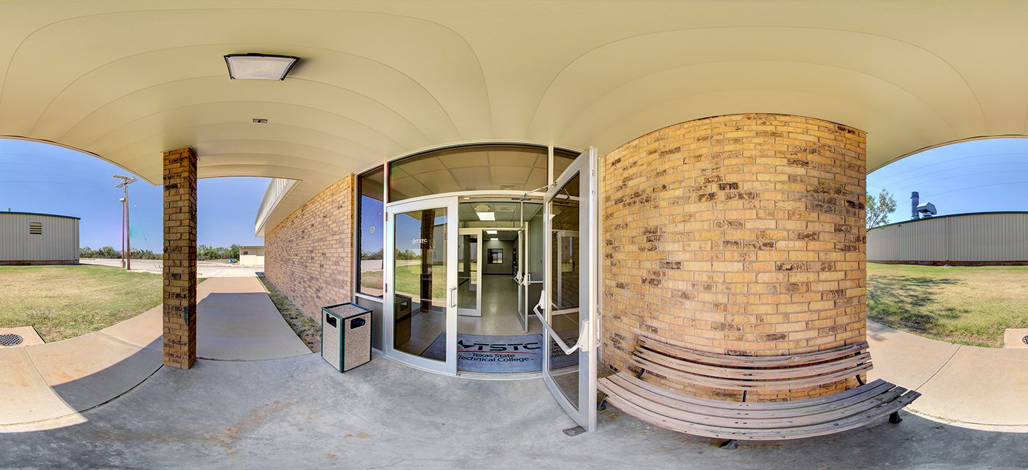 Panoramic view of a brick building entrance with glass double doors labeled Texas State Technical College, featuring a covered walkway, bench seating, and adjacent grassy areas outside.