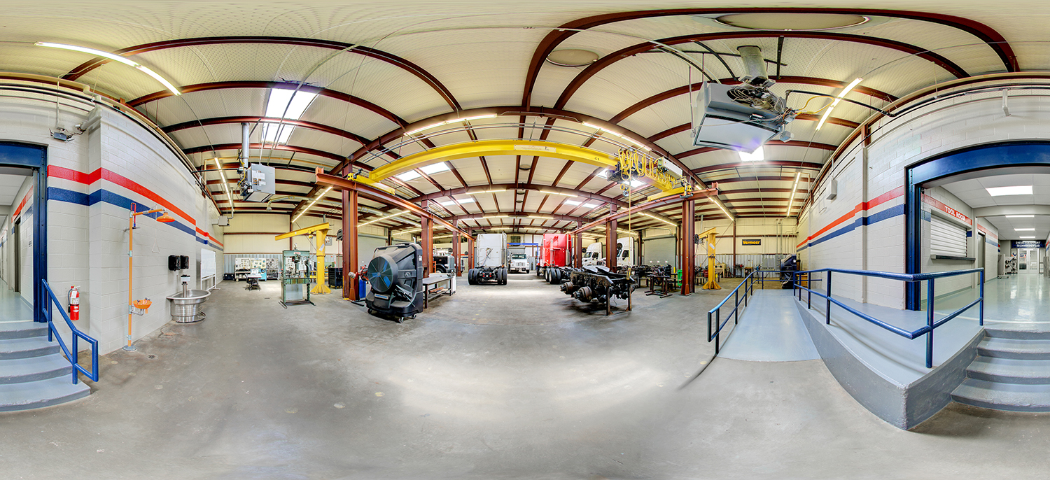 Panoramic view of an automotive and diesel training lab with large trucks, exposed chassis components, overhead cranes, and workshop equipment inside a high-ceiling industrial space.