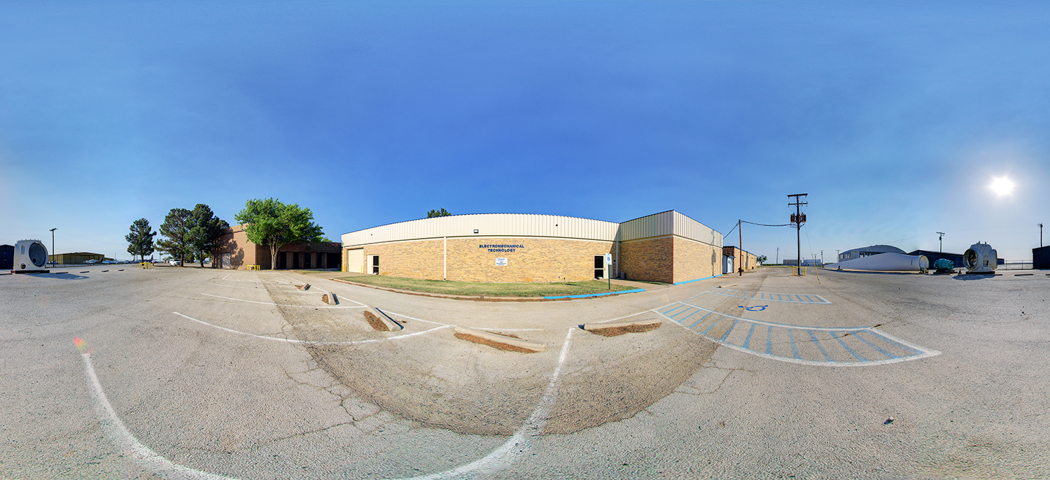 Panoramic view of a low brick building labeled “Electromechanical Technology,” surrounded by a wide parking lot with marked spaces, industrial equipment nearby, and a clear sunny sky overhead.