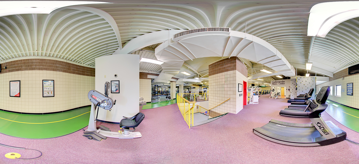 Panoramic view of a fitness center with cardio equipment including treadmills and exercise machines, a walking track, weight training areas, and a central staircase under a high, curved ceiling with exposed beams.
