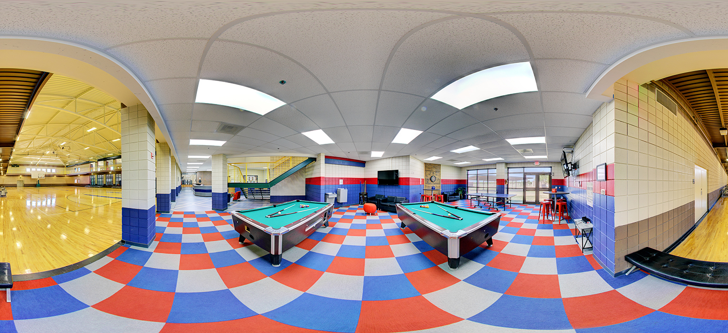 Panoramic view of a recreation area next to a gymnasium, featuring pool tables on a red, blue, and gray checkered floor, seating and tables along the walls, and an adjacent basketball court visible through wide openings.