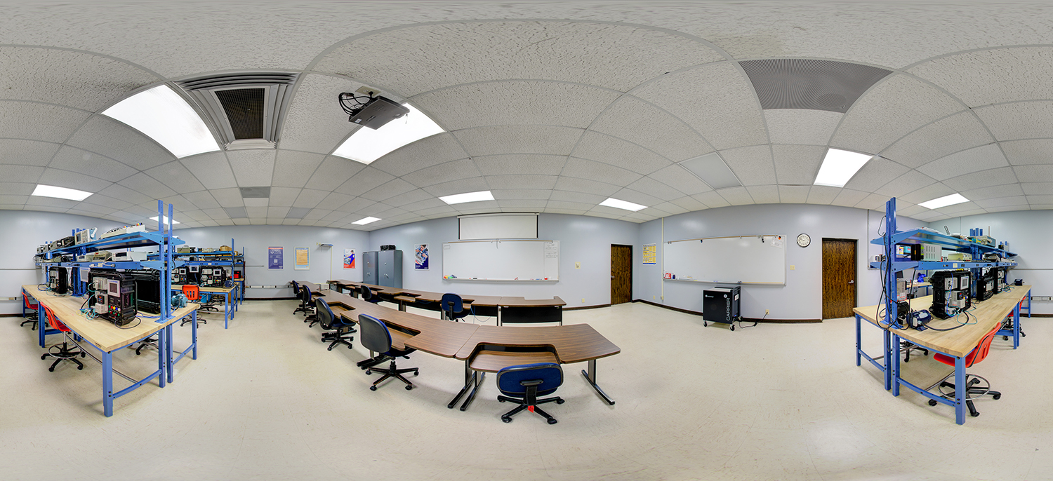 Panoramic view of an electronics classroom with workbenches holding testing equipment, rows of desks and chairs, whiteboards at the front, and instructional posters on the walls.