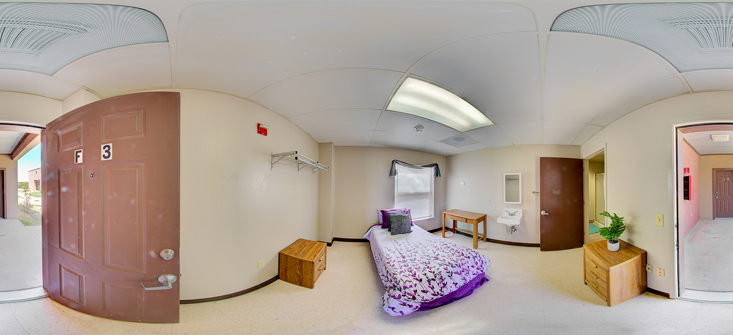 Panoramic view of a small dorm-style room with a single bed dressed in purple bedding, wooden furniture including a dresser and desk, a wall-mounted sink with mirror, and an open door labeled “F3” leading to an outdoor walkway.