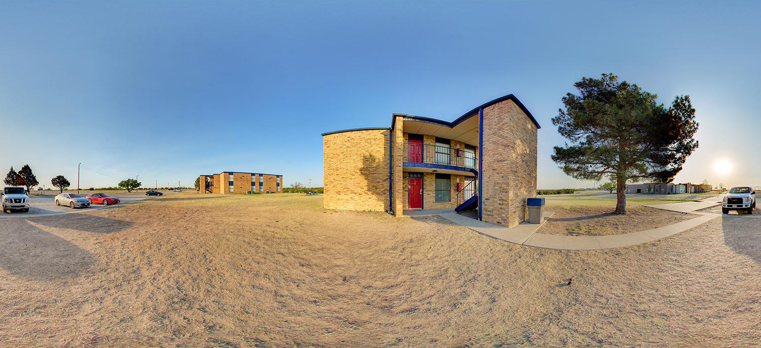 Panoramic view of a small two-story brick residential-style building with exterior stairs and red doors, surrounded by a dry grassy area, parked cars, and a few trees under a clear sky.