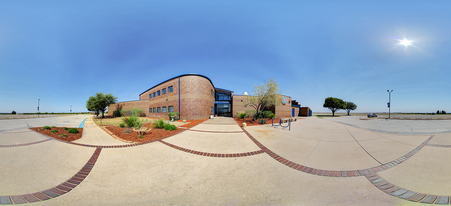 Panoramic view of a modern campus building with a curved brick facade and glass entrance, surrounded by landscaped walkways, small trees, and an open parking area under a bright, cloudless sky.