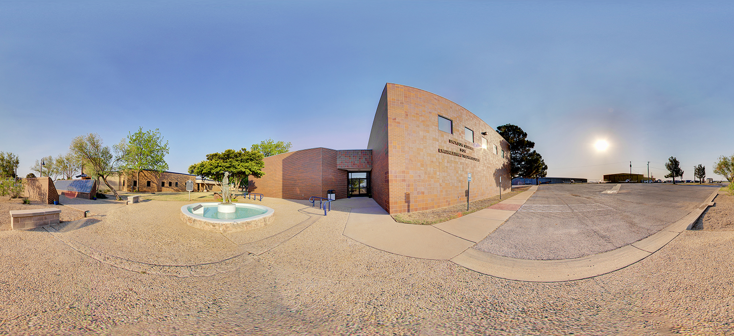 Panoramic view of an engineering building exterior with a curved brick façade, a small entrance, and a courtyard featuring a fountain, benches, and desert landscaping under a bright sky.