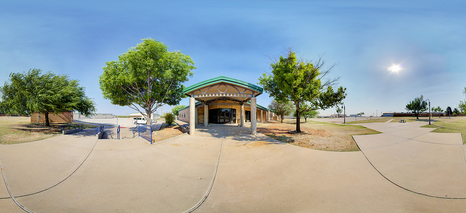 Panoramic view of a campus building entrance with a covered portico supported by columns, surrounded by trees and walkways under a bright, clear sky, with open grounds and pathways extending in multiple directions.