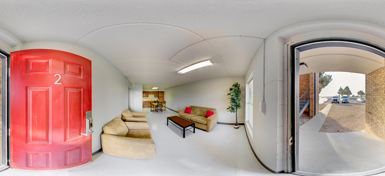 Panoramic view of a small apartment-style living room with two beige sofas, a coffee table, and a dining area leading to a compact kitchen; a bright red door labeled “2” stands open to an exterior walkway with parked cars visible outside.