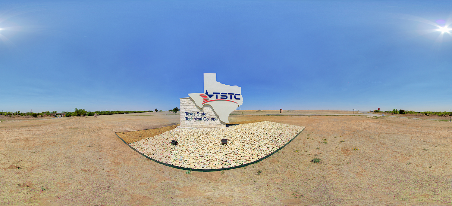 Panoramic view of a roadside sign shaped like the state of Texas displaying “TSTC – Texas State Technical College,” set on a bed of rocks in a dry, open landscape under a bright, clear sky.