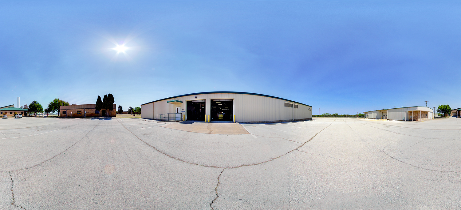 Panoramic view of a large industrial training building with wide garage-style doors open to a spacious parking lot, surrounded by additional low buildings under a bright midday sky.
