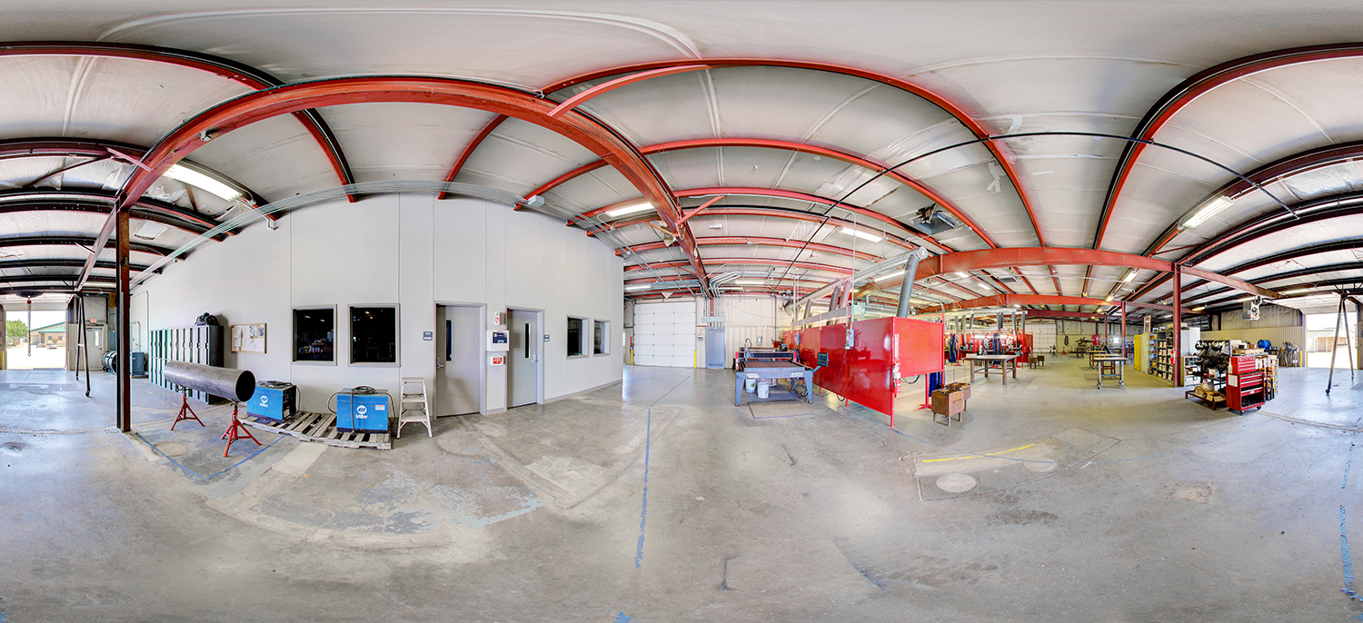 Panoramic view of a welding and fabrication lab with red steel beams overhead, welding booths with red safety curtains, metal worktables, tool chests, and industrial equipment arranged across a spacious concrete floor.