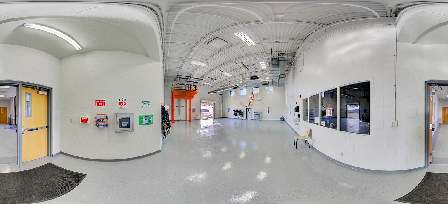 Panoramic view of a spacious industrial lab with white walls, polished floors, and exposed ceiling ducts, featuring safety equipment on one wall, large windows into an adjacent room, and an open garage-style door letting in daylight.
