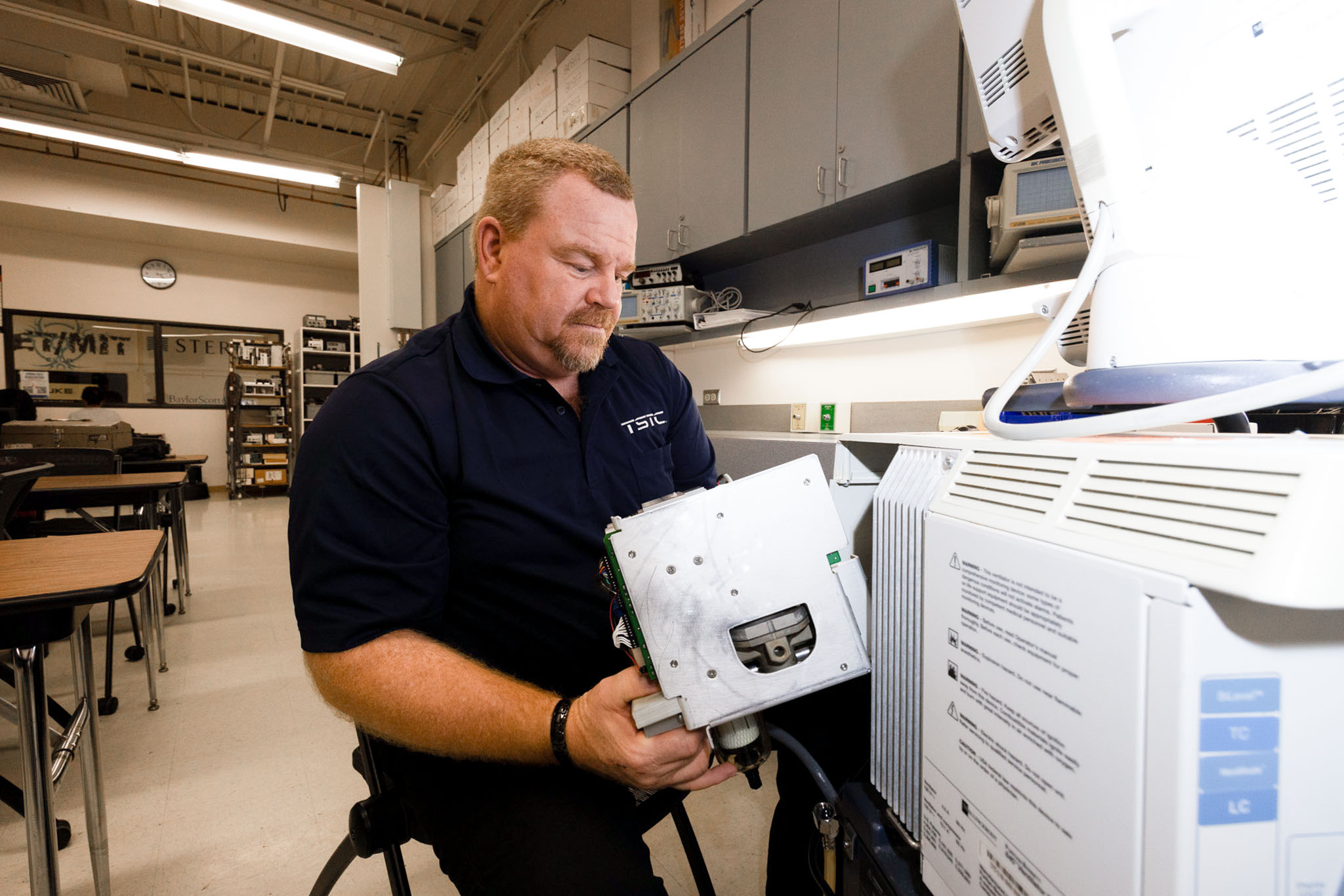 Man in a lab classroom installs or removes an internal electronic component from a medical machine, with testing equipment and storage cabinets in the background.