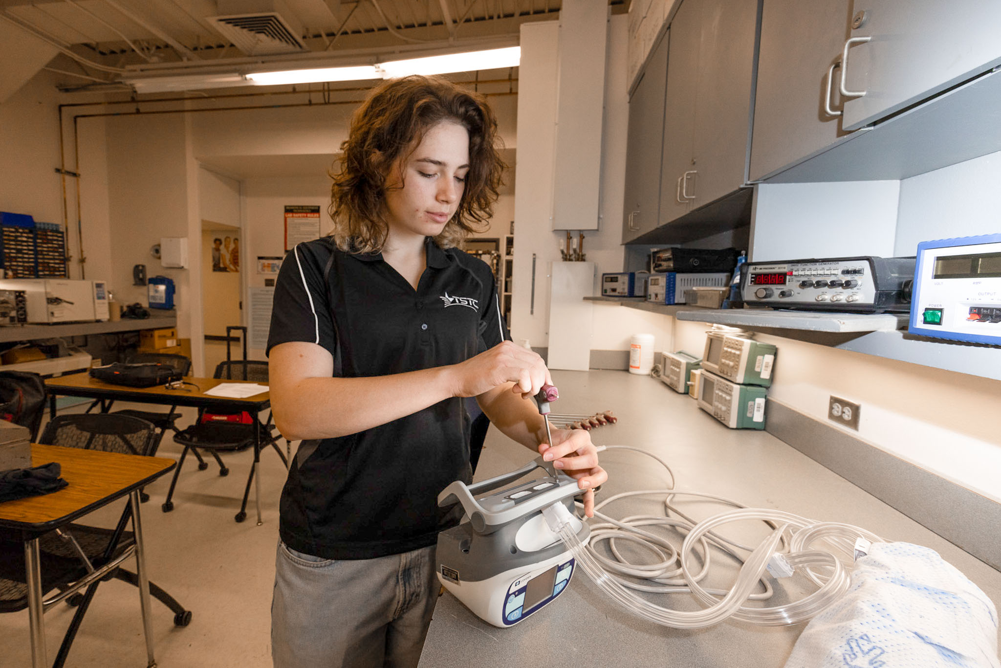 Young woman in a technical lab uses a screwdriver to adjust or repair a medical device with attached tubing, standing at a workbench surrounded by testing equipment.
