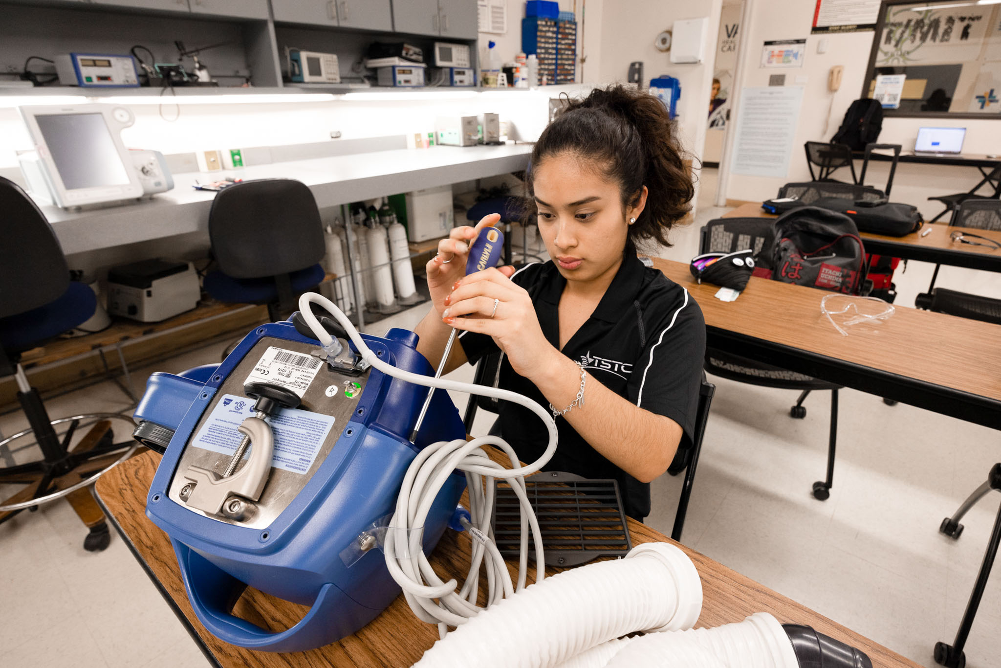 A student in a lab concentrates while using a screwdriver to adjust a blue medical device with coiled tubing, seated at a classroom workbench with testing equipment in the background.
