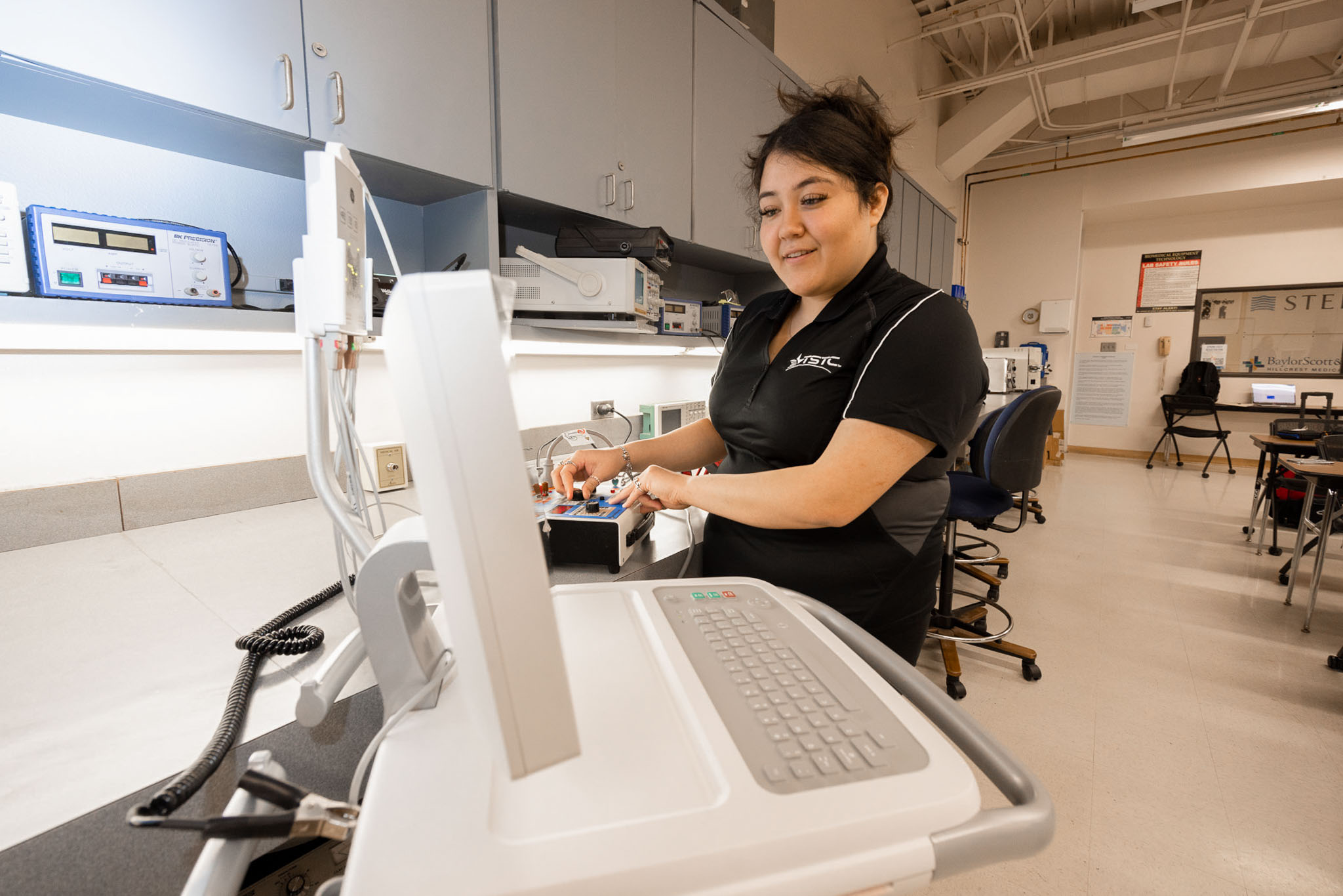 Woman in a clinical training lab operates and tests medical equipment on a wheeled cart, adjusting controls while surrounded by diagnostic devices and classroom workstations.
