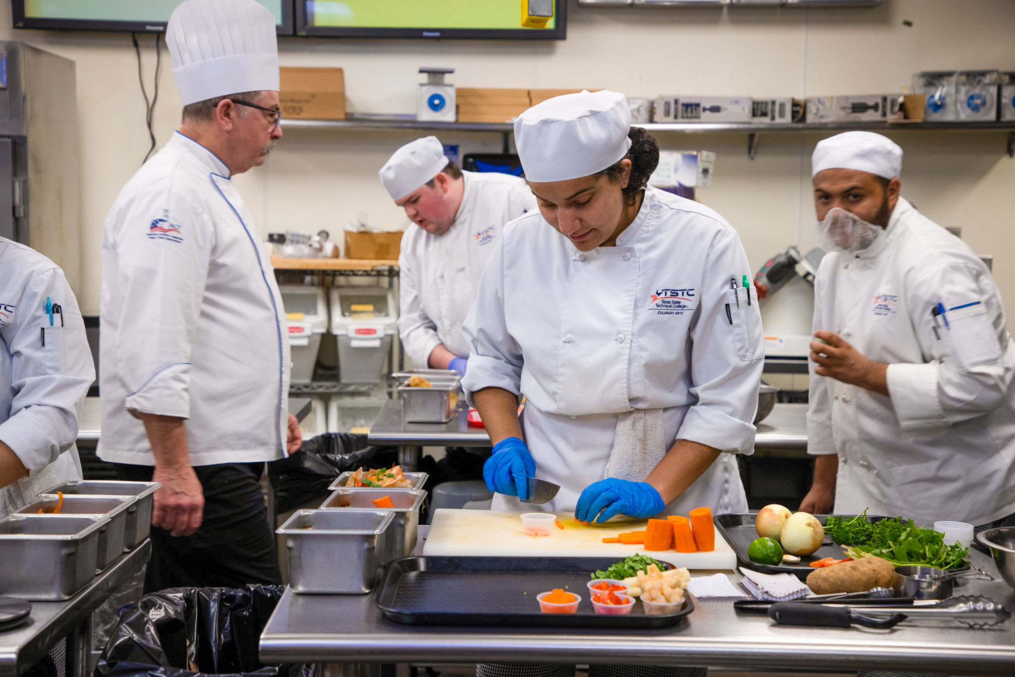Culinary students in white chef uniforms and hats prepare food in a commercial kitchen, with one student chopping carrots on a cutting board while others work behind them.