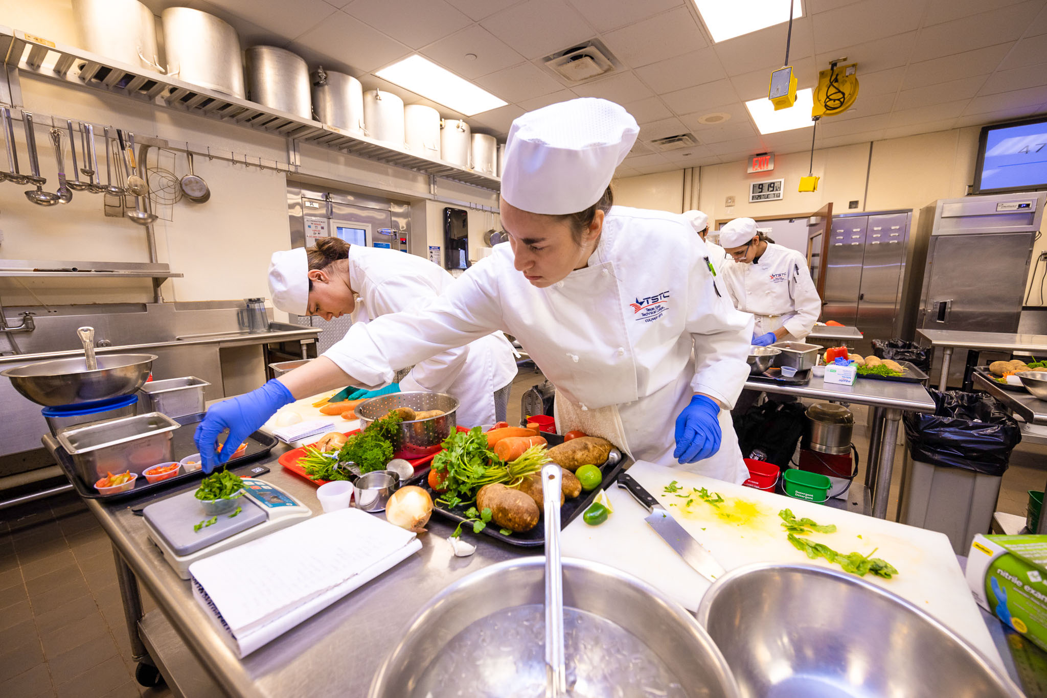Culinary student in a white chef uniform and hat, wearing blue gloves, reaches across a stainless steel workstation to weigh chopped herbs, with fresh vegetables and other students preparing ingredients in a commercial kitchen.