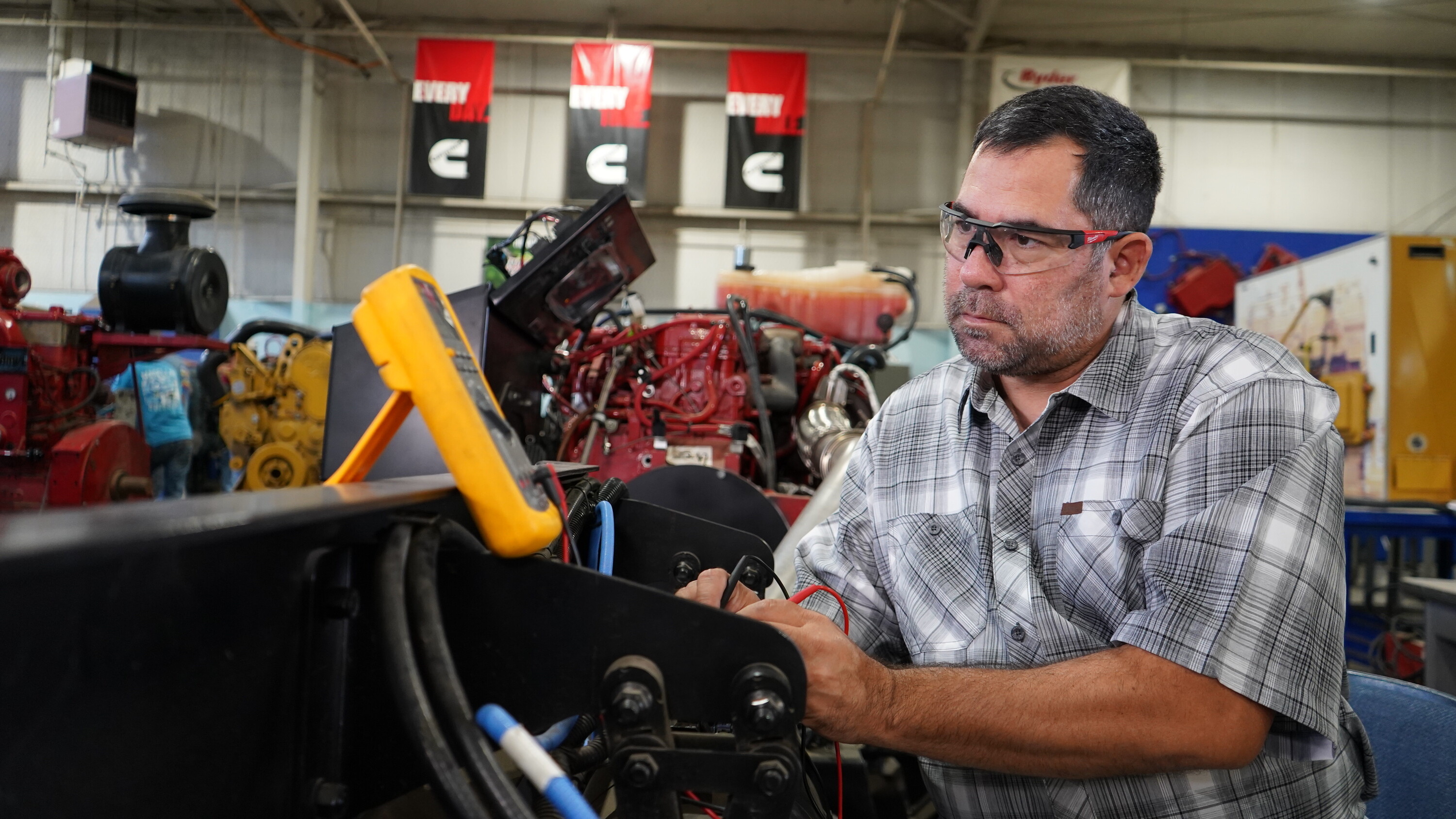 A man wearing safety glasses uses a handheld multimeter to test electrical components on a machine, concentrating as he works in a workshop filled with engines and industrial equipment.