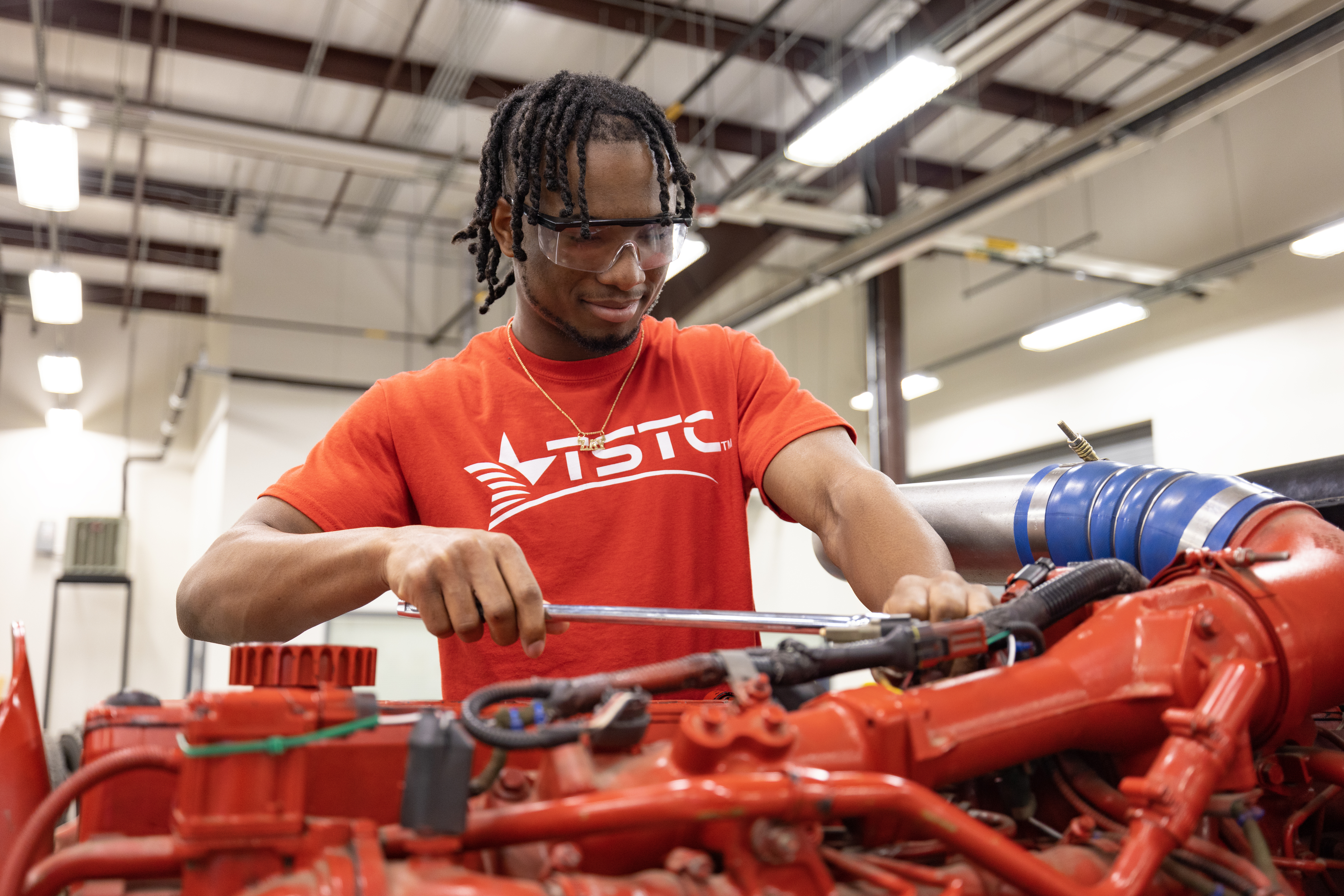 A young man wearing safety glasses and a red TSTC shirt uses a wrench to work on a large red engine, focused as he tightens a component in a workshop.