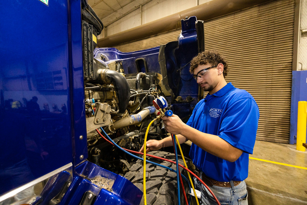 A student wearing safety glasses and a blue TSTC polo connects diagnostic hoses to a large truck engine, checking systems inside a service bay with the hood open.