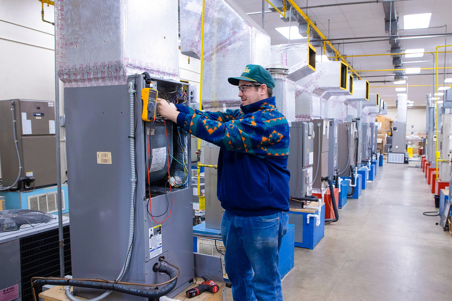 A student wearing safety glasses and a cap uses a multimeter to test wiring inside an open HVAC unit, standing in a lab lined with similar systems and ventilation ductwork.