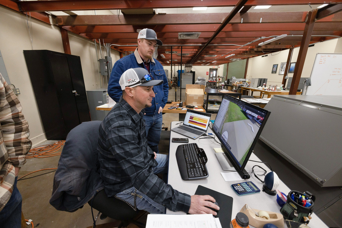 Two students in an HVAC lab review data on a desktop monitor, one seated using a mouse and keyboard while the other stands behind observing, with tools, laptops, and equipment spread across the workspace.