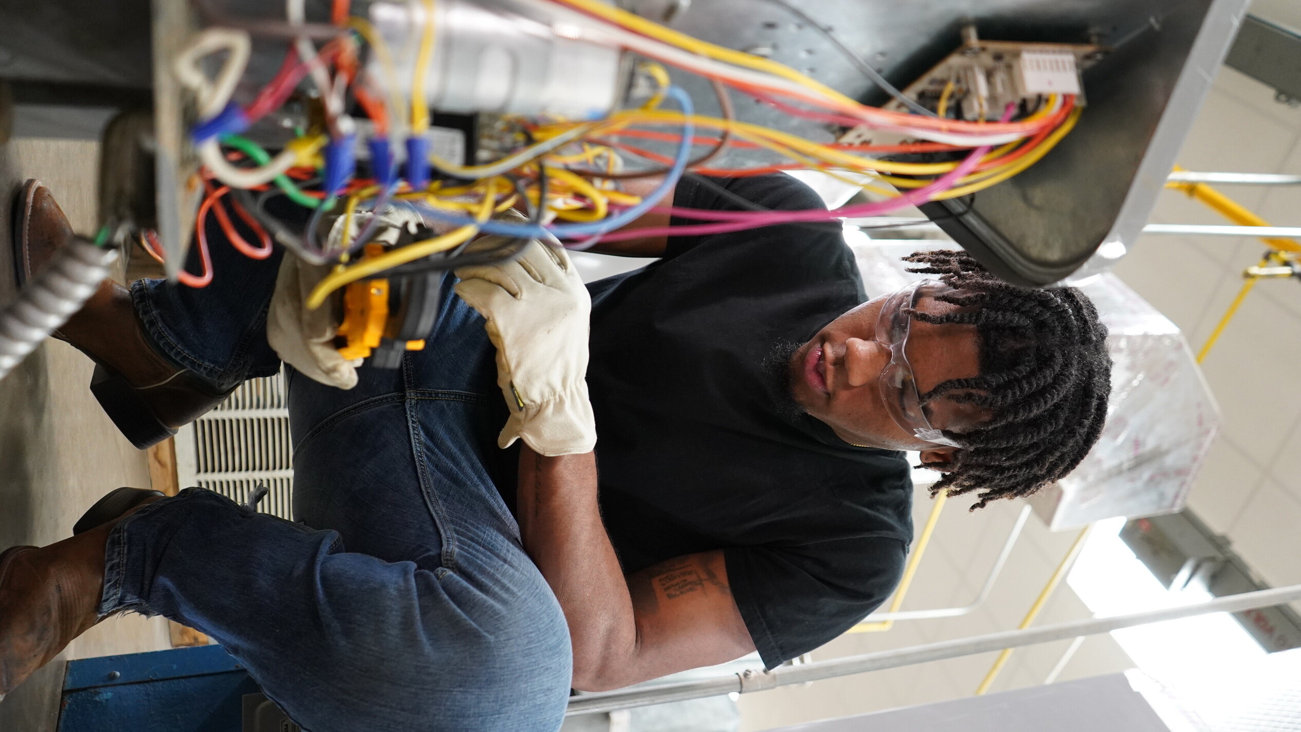 A student wearing safety glasses and gloves crouches while using a multimeter to test colorful wiring inside an open HVAC unit, focusing intently on the electrical components.