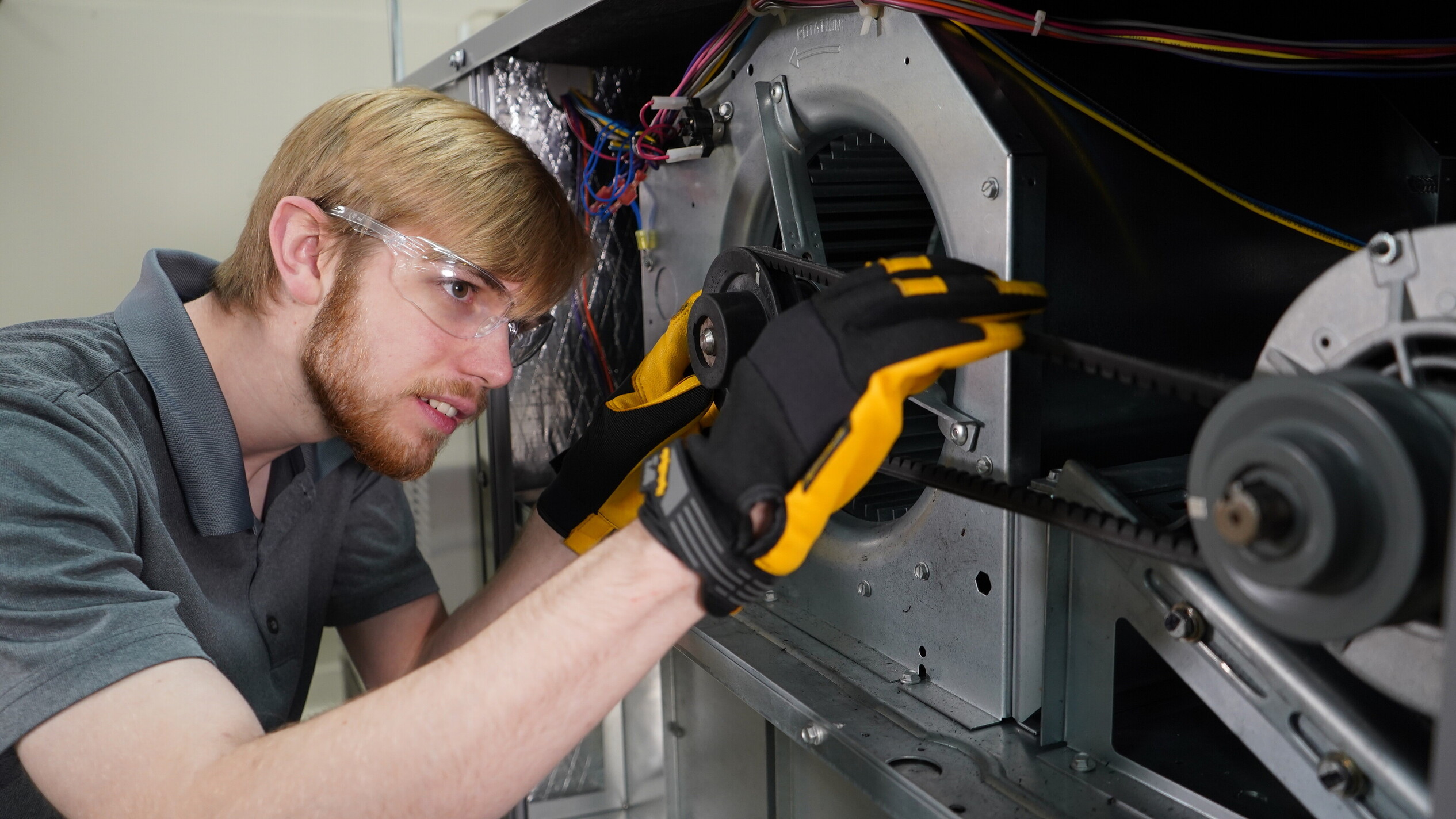 A student wearing safety glasses and protective gloves adjusts a belt inside an open HVAC unit, carefully aligning the pulley system while inspecting internal components.