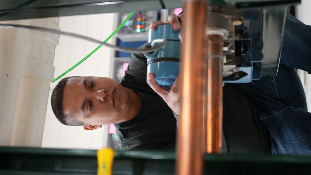 A student crouches in a workshop, concentrating as he adjusts a blue mechanical component connected to copper piping and wiring, with industrial equipment in the background.