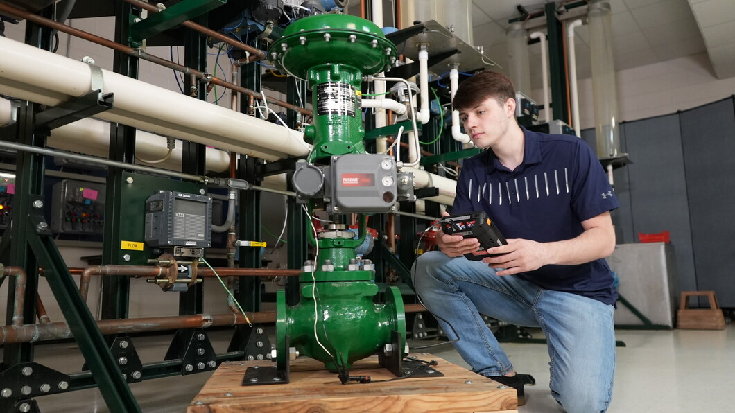 Student kneels beside an industrial piping system, using a handheld device to test a large green valve with connected sensors and wiring.