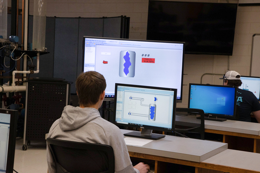Student sits at a workstation, viewing a computer simulation of a component on dual monitors in a technical lab setting.