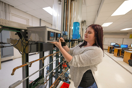 Student stands at an industrial training rig, adjusting a digital control panel connected to copper piping and fluid system components in a lab.