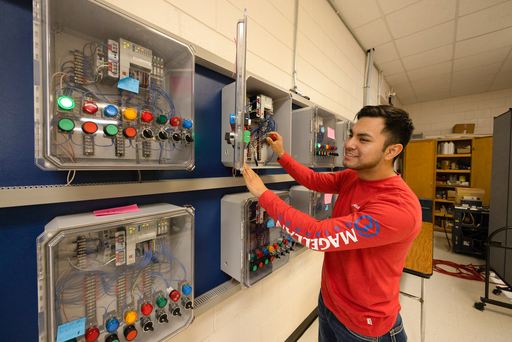 Student stands at an electrical training panel, adjusting control switches and wiring inside an open control box in a lab setting.