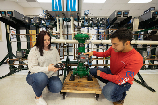Students kneel beside an industrial piping system, using handheld devices to test a large green valve with connected sensors and control components in a lab.