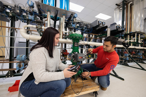 Students kneel beside an industrial piping system, using handheld devices to measure and adjust a green control valve with connected sensors and wiring in a lab.
