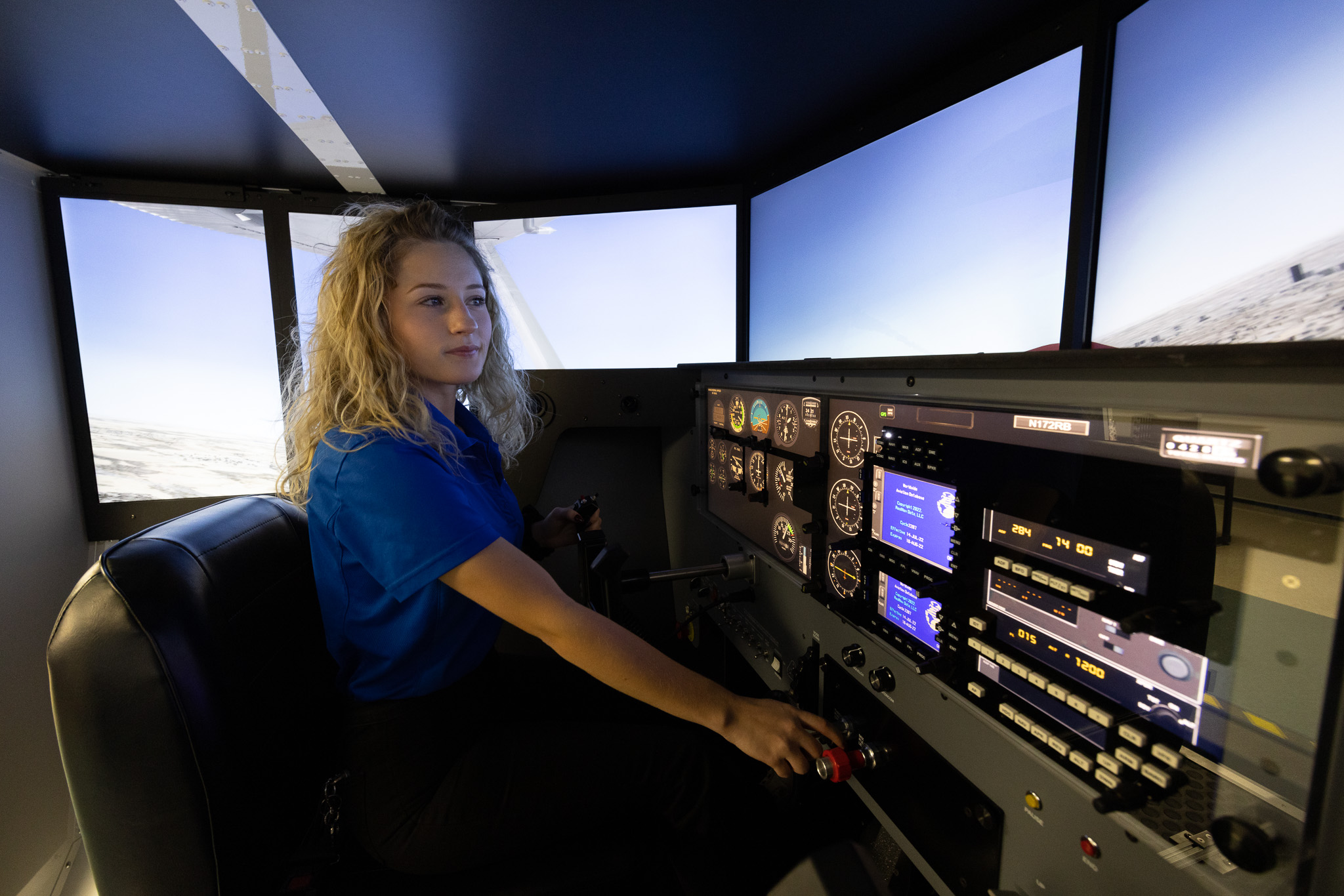 Student sits in a flight simulator, operating cockpit controls while viewing a simulated flight environment on surrounding screens.