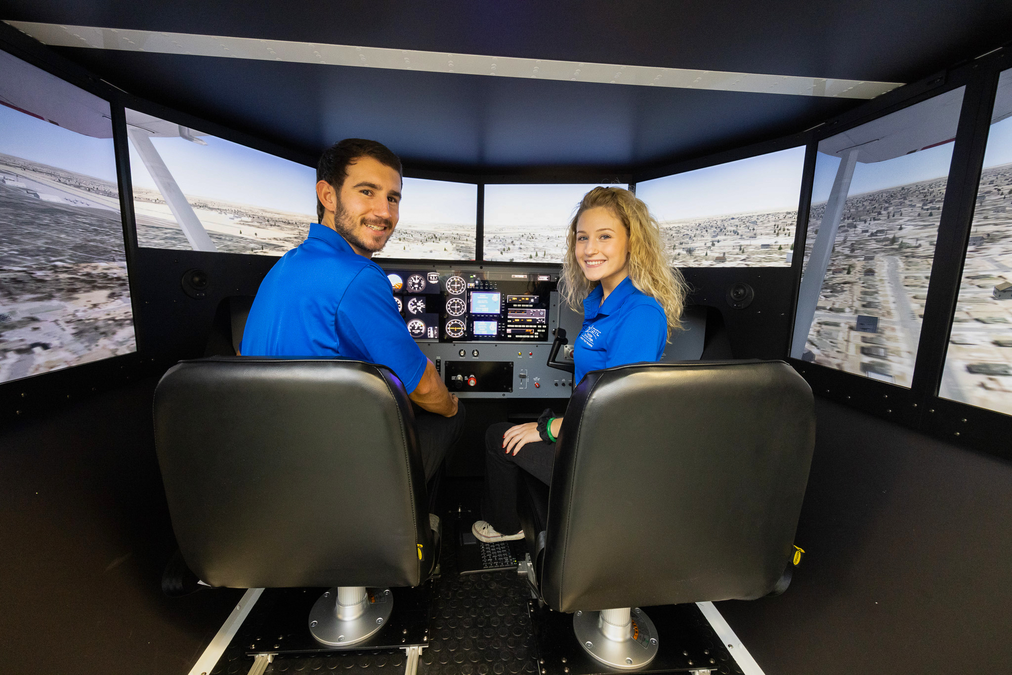 Two students sit in a flight simulator cockpit, operating controls while a simulated landscape is displayed across surrounding screens.