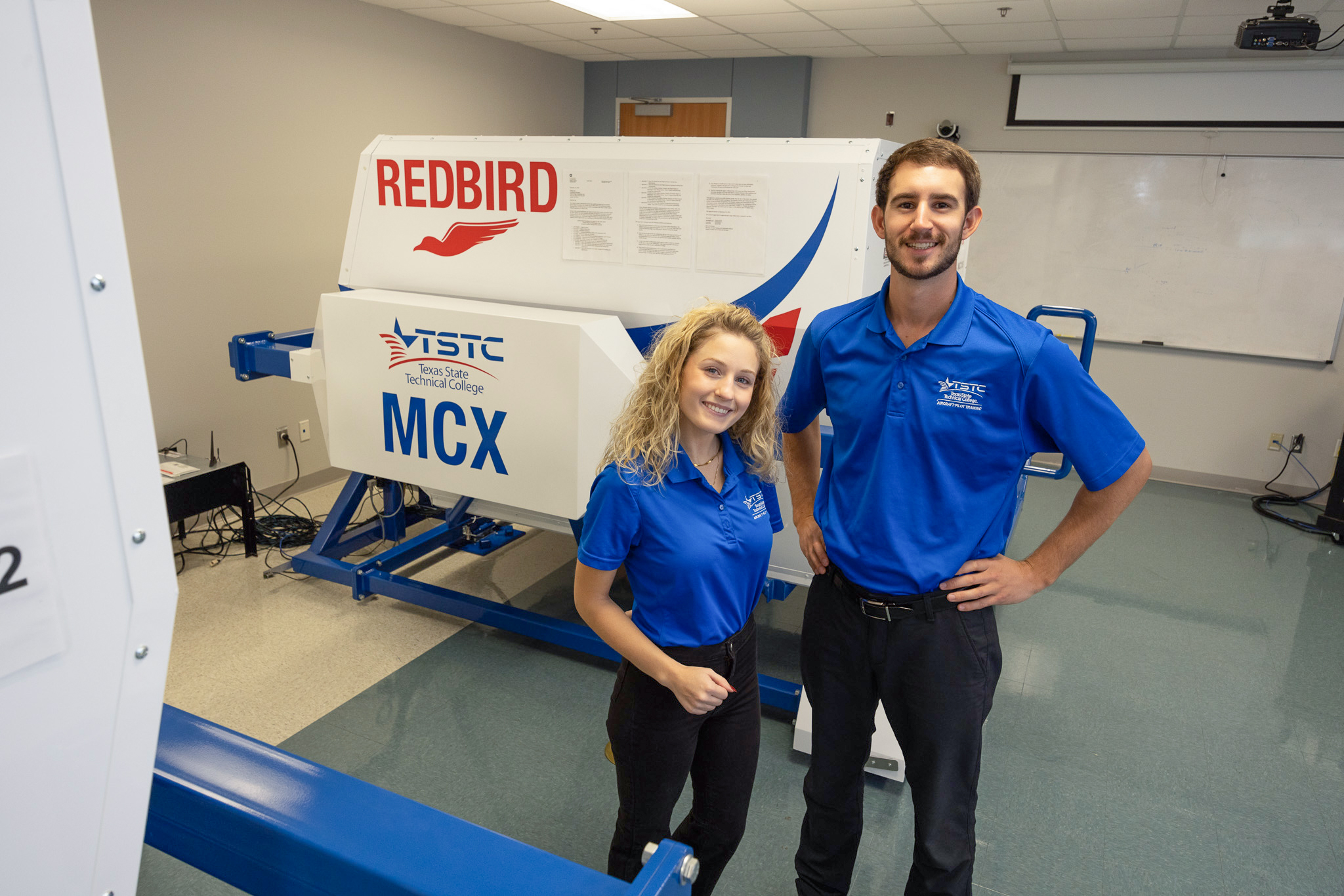 Two students stand beside a Redbird flight simulator in a training lab, posing in front of the equipment.