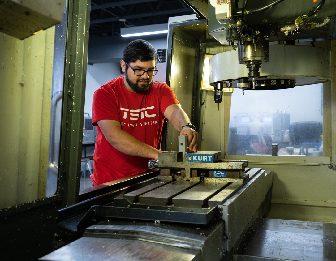 A student wearing glasses and a red shirt sets up a metal workpiece in a vise inside a CNC milling machine, carefully positioning it beneath the cutting spindle in a workshop.