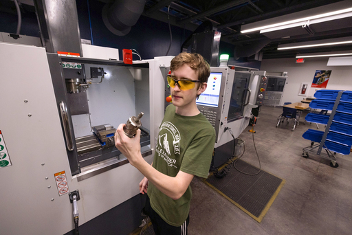 A student wearing yellow safety glasses stands at an open CNC machine, holding and inspecting a metal tool or part in a manufacturing lab.
