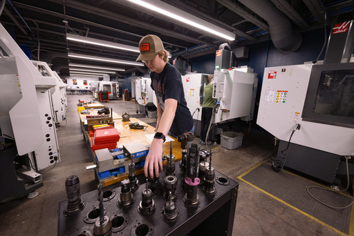 A student in a cap reaches for cutting tools organized in a holder, selecting a tool in a CNC machining lab with multiple machines in the background.