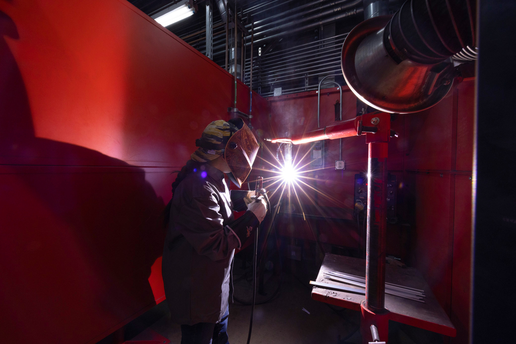 A student wearing a welding helmet and protective jacket welds a metal piece inside a red booth, producing an intense bright arc and sparks, with ventilation equipment and tools nearby.